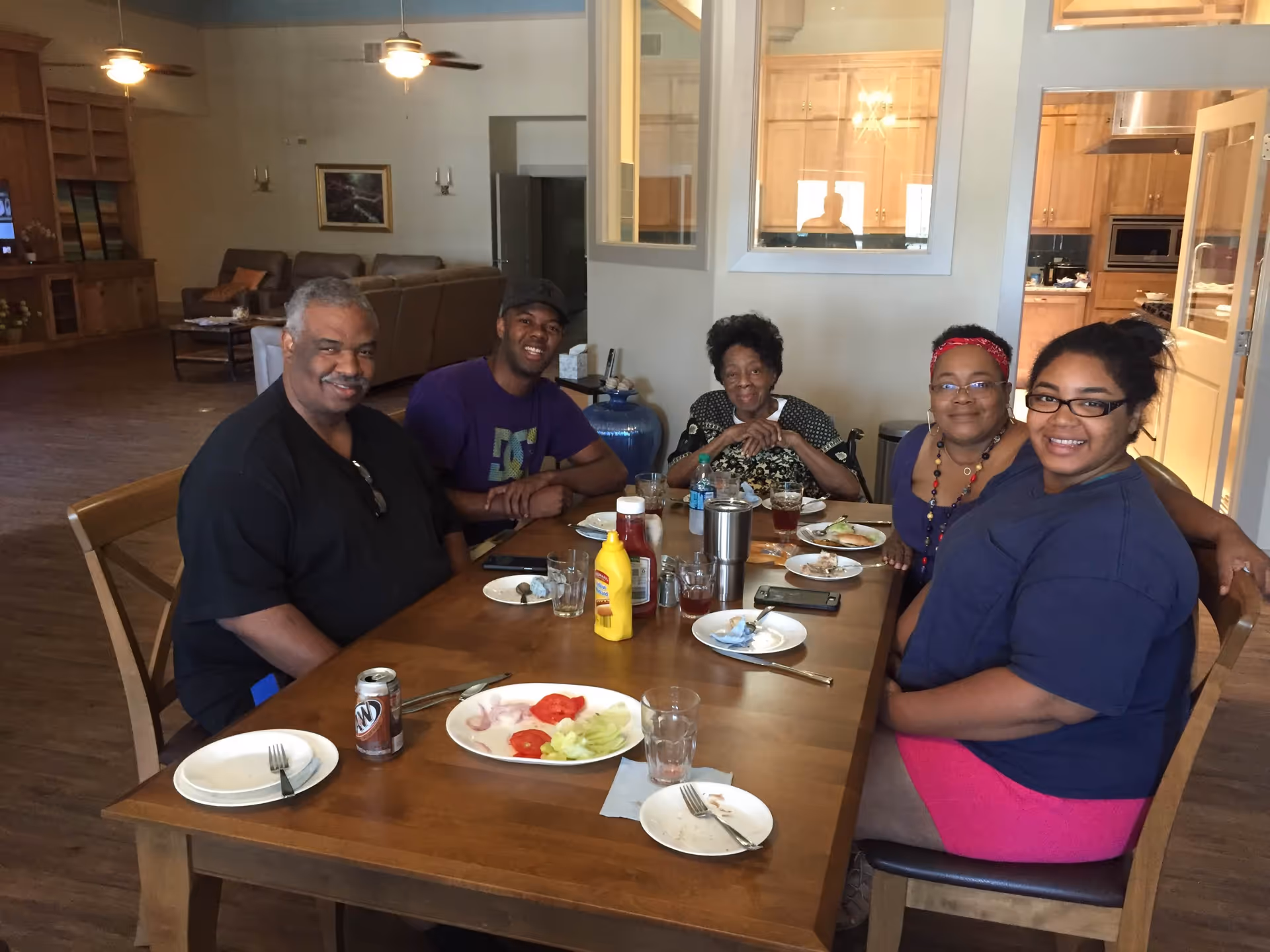 Five people sitting around a wooden dining table with plates of food and drinks, smiling at the camera in a well-lit room with ceiling fans, a couch, and a kitchen visible in the background.