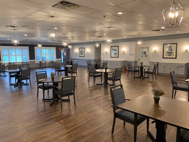 A spacious dining room with multiple round and square tables, each surrounded by chairs. The room has wooden flooring, soft lighting from hanging lantern-style fixtures, and framed artwork on the walls. Large windows with white shutters allow natural light to enter the space.
