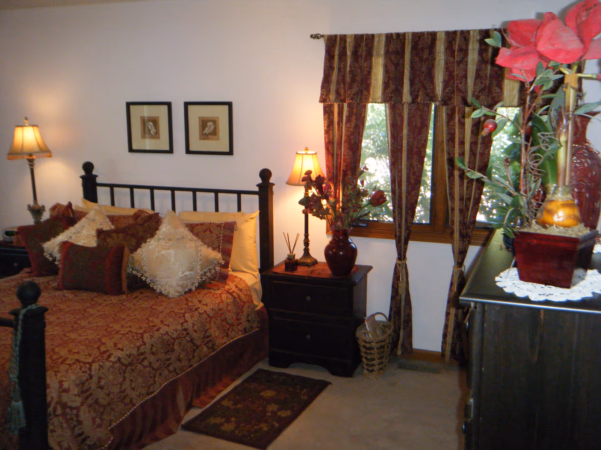 A cozy bedroom with a dark wooden bed frame and a bed covered in a patterned red and gold bedspread with multiple decorative pillows. There are two framed pictures on the wall above the bed. On either side of the bed are dark wooden nightstands with lamps and decorative items. A window with red and gold striped curtains is on the right side of the room, and a dresser with floral decorations is partially visible in the foreground.