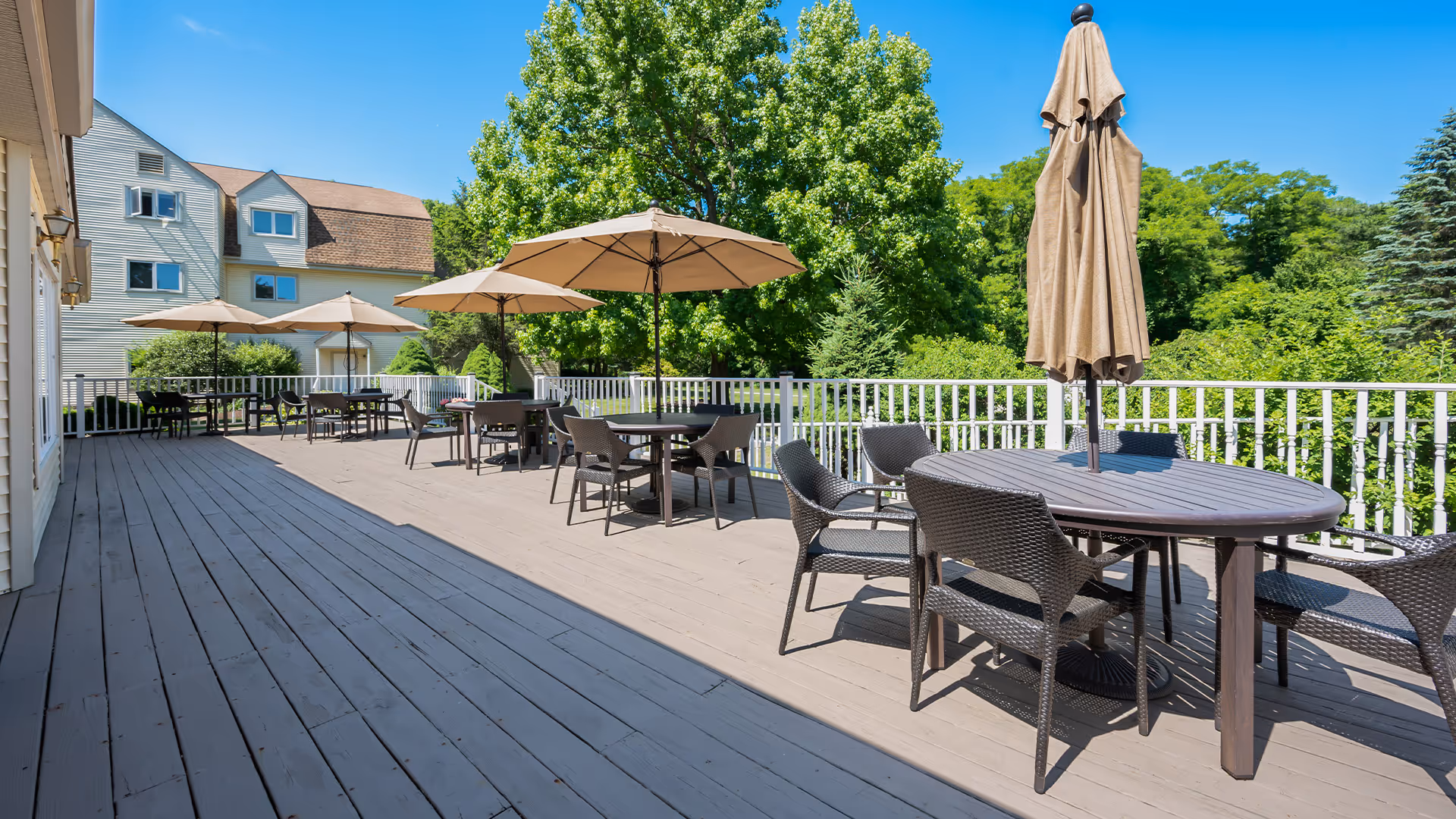 Spacious outdoor deck with round tables, wicker chairs and umbrellas overlooking trees and a nearby building.