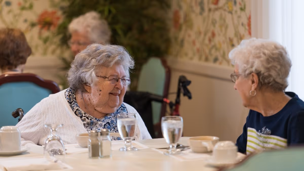 Two elderly women smiling and chatting while seated at a dining table with glasses and bowls in a senior living facility dining room.