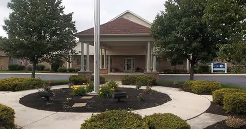 Covered entrance of a single-story brick retirement community building with a porte-cochère, flagpole and circular landscaped drive.