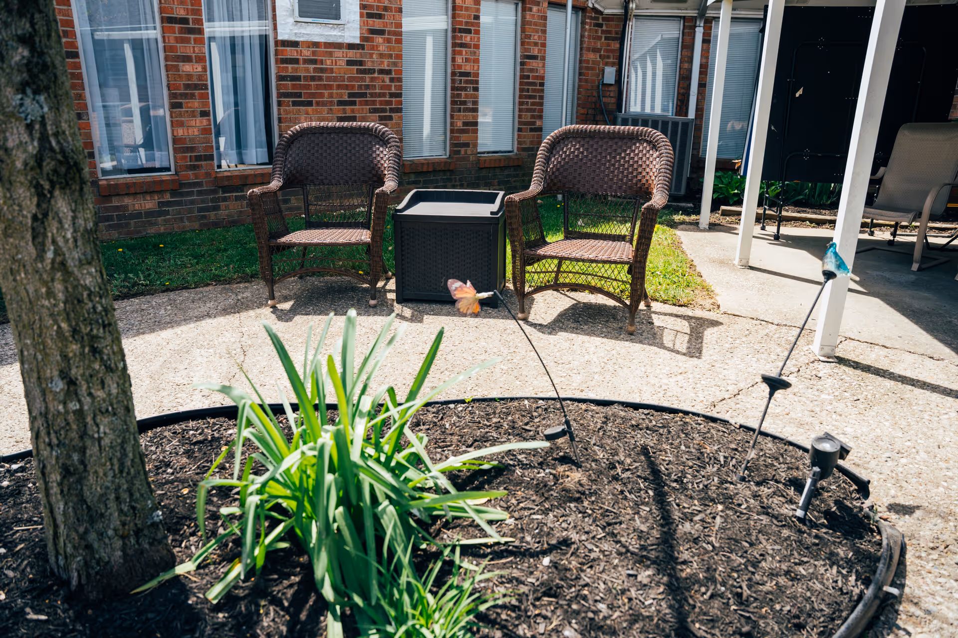 Outdoor patio area with two wicker chairs and a small table between them, situated on a concrete surface next to a garden bed with green plants and mulch. The background shows a brick building with several windows and a covered area with additional seating.