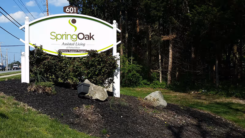 Roadside sign for Spring Oak Assisted Living of Forked River in a landscaped bed beside a road and woods.