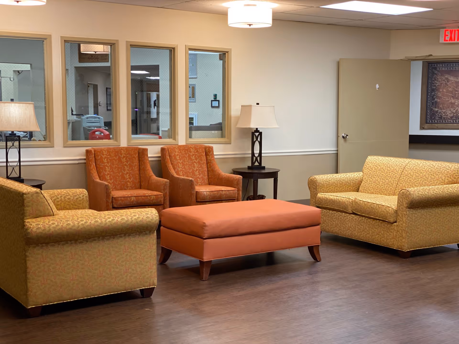 A cozy seating area in a senior living facility with two patterned yellow sofas, two orange armchairs, and a large orange ottoman in the center. There are two side tables with lamps on either side of the armchairs. The walls are beige with three windows showing an interior hallway, and a door is open to the right. The floor is wooden.