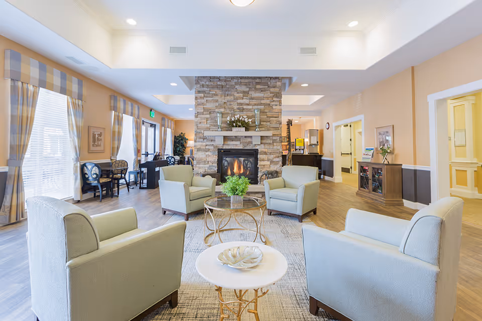 A bright and spacious living room area in an assisted living facility featuring four light green armchairs arranged around two small round tables. A stone fireplace with a mantle decorated with vases and flowers is centered in the background. Large windows with plaid curtains allow natural light to fill the room, and additional seating is visible near the windows. The room has wood flooring and soft beige walls with white trim.