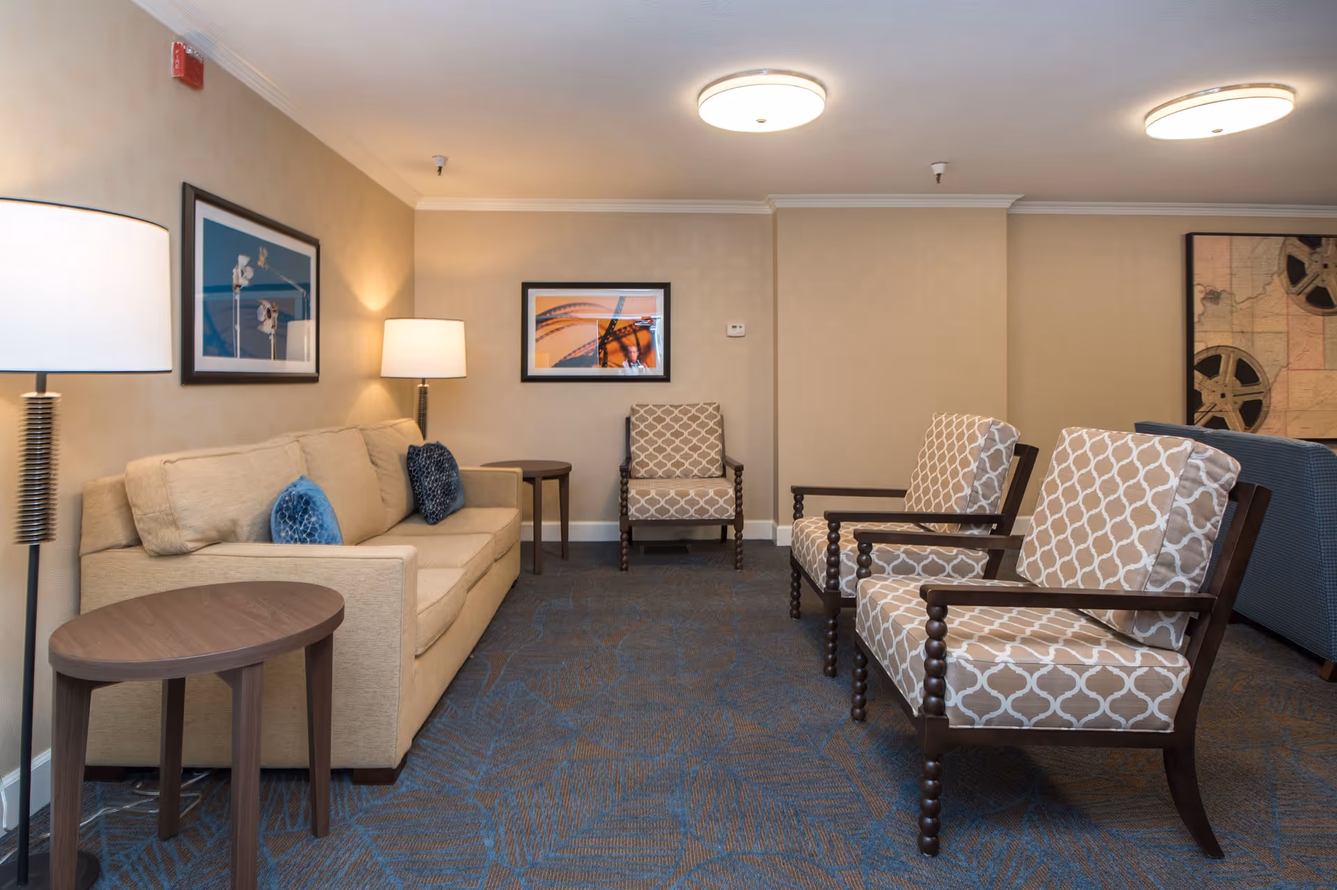 A cozy living room area with a beige sofa adorned with blue and beige cushions, two patterned armchairs with wooden frames, a small round wooden side table, and two floor lamps. The walls are decorated with framed artwork, and the carpet has a blue and brown geometric pattern.
