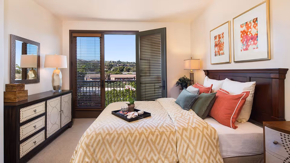A well-lit bedroom with a large bed featuring multiple pillows in red, green, and white. The bed has a beige patterned blanket. There is a wooden headboard and two framed abstract artworks above it. To the left, there is a dresser with drawers and a lamp, along with a mirror on the wall. A door with glass panels and blinds opens to a balcony with a view of greenery and distant buildings.