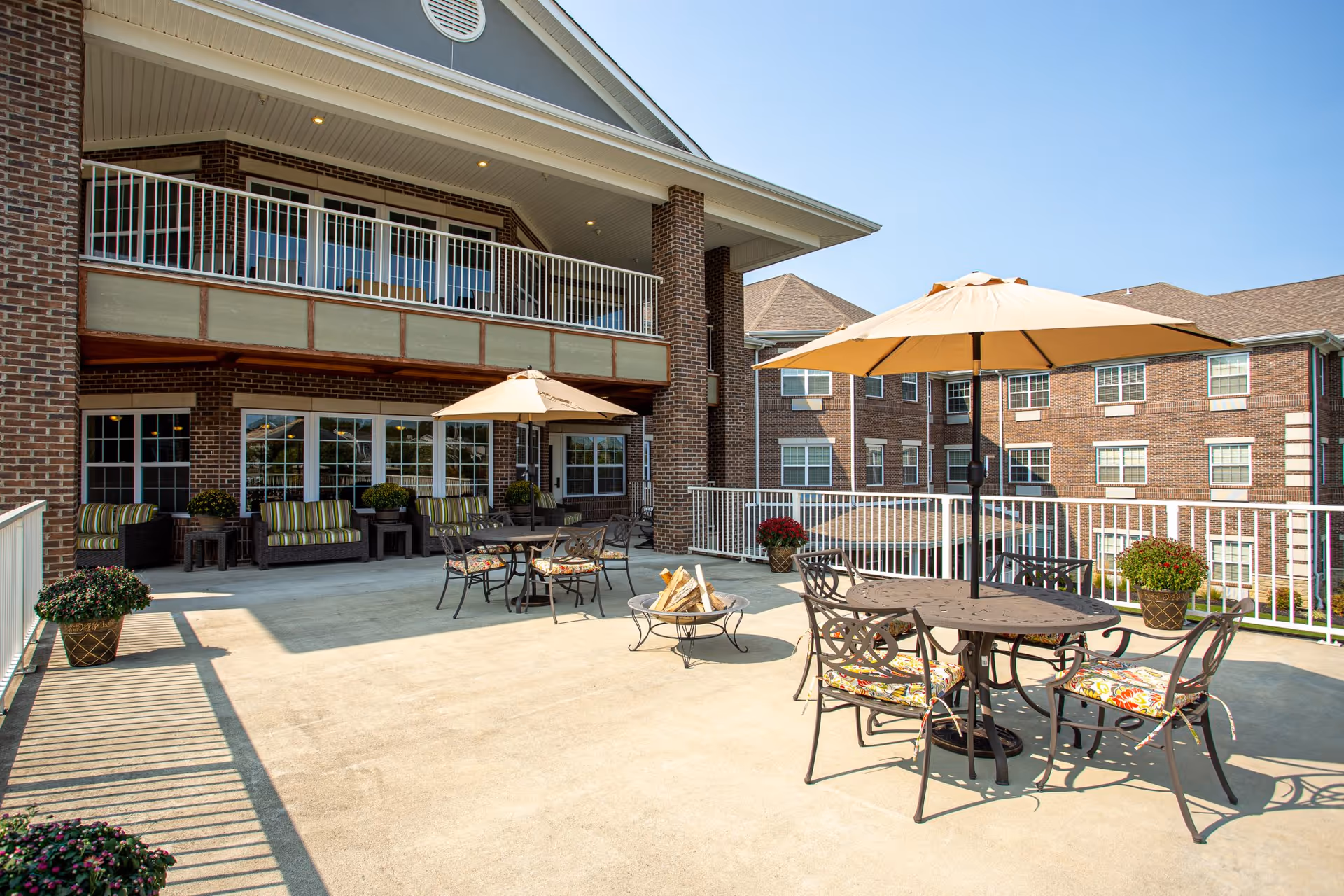 Outdoor patio area at Magnolia Springs Florence with metal tables and chairs featuring floral cushions, large beige umbrellas, potted plants, and a fire pit. The patio is adjacent to a brick building with multiple windows and a balcony above.