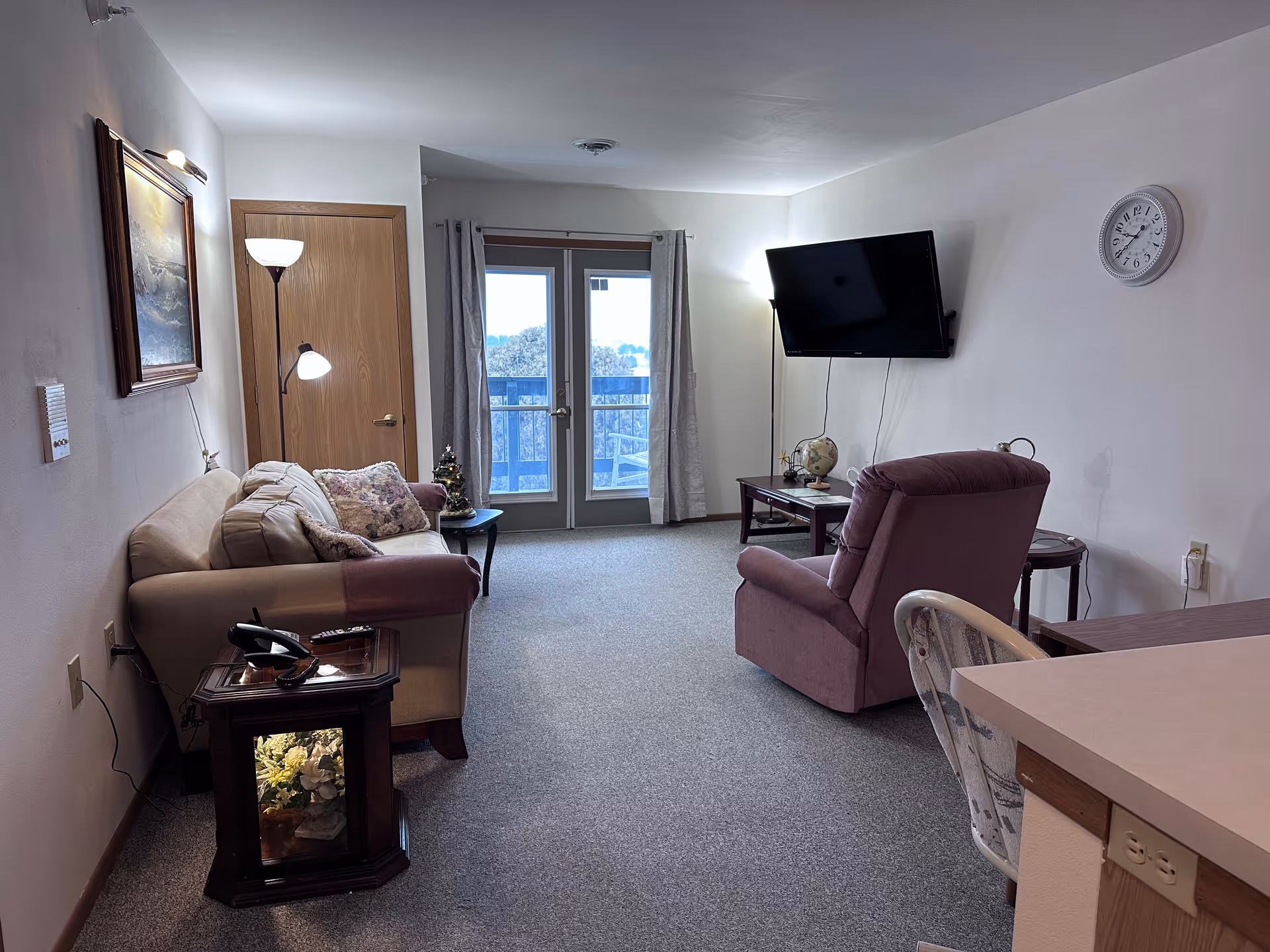 A living room with a beige sofa and a purple armchair facing a wall-mounted flat-screen TV. There is a small wooden side table with a lamp and a globe, a floor lamp, a wall clock, and a painting on the wall. French doors with curtains lead to a balcony with outdoor chairs visible.