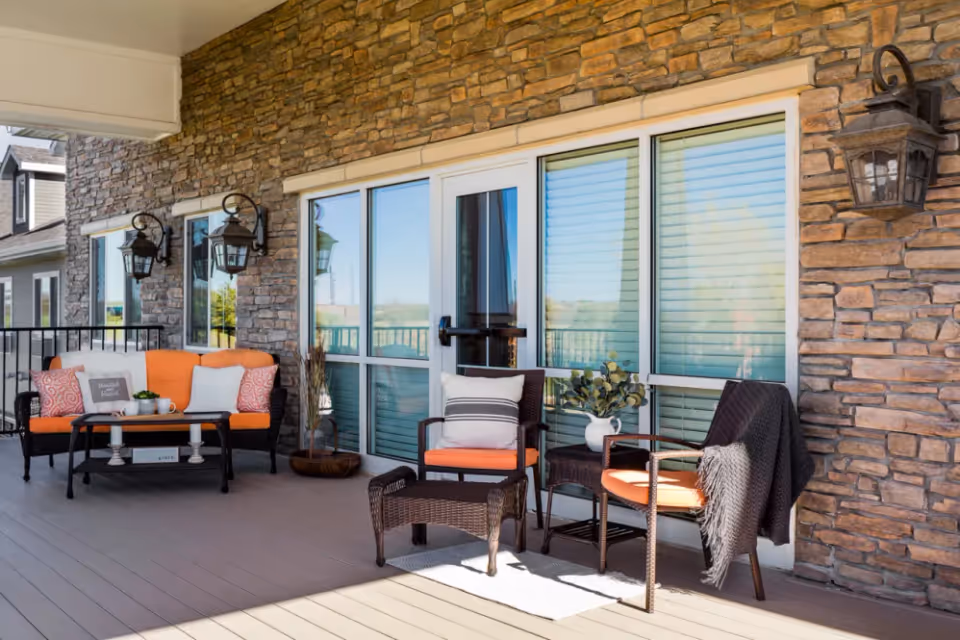 Outdoor patio area with stone wall and large windows. The patio features a black wicker loveseat with orange cushions and decorative pillows, a black coffee table with a small plant and a cup, two black wicker chairs with orange cushions, one with a striped pillow and the other draped with a black blanket, and a small black wicker footrest on a light-colored rug. There are three black wall-mounted lantern-style lights on the stone wall.