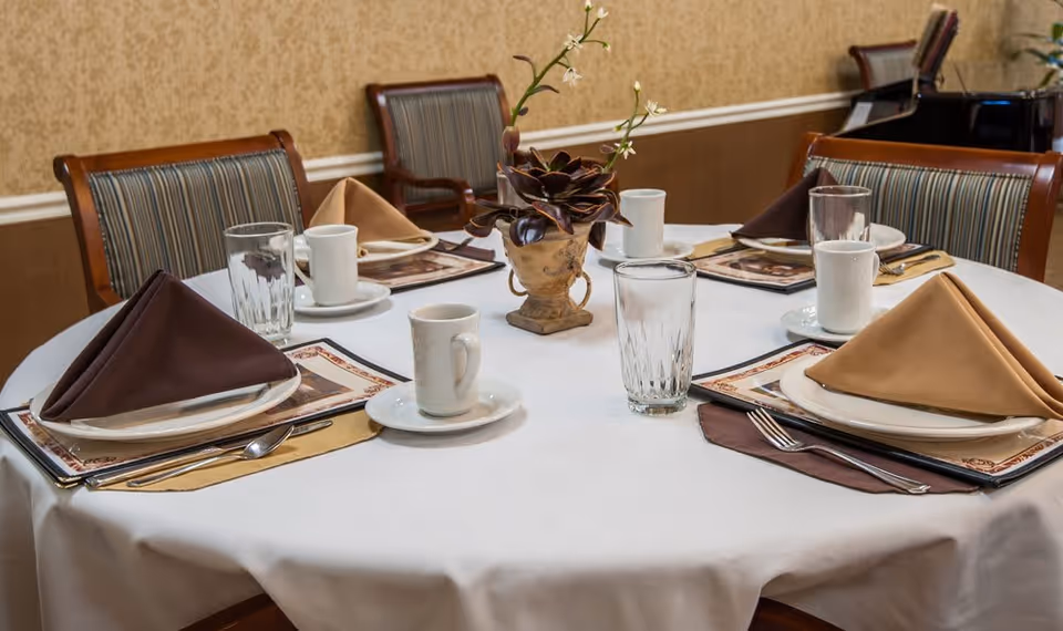 A round dining table set for four with white tablecloth, plates, cups, glasses, silverware, and folded napkins in brown and beige colors. A small decorative plant is placed in the center of the table. The chairs around the table have wooden frames and striped upholstery. The background shows a beige patterned wall.