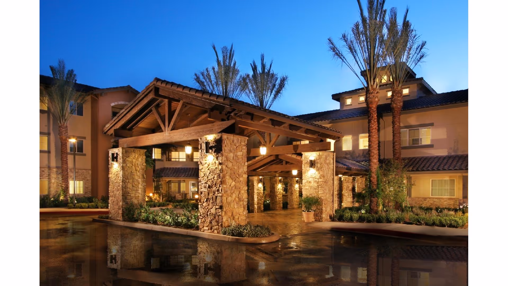 Exterior view of Valencia Terrace senior living facility at dusk, featuring a covered entrance with stone pillars, warm lighting, palm trees, and a multi-story building in the background.