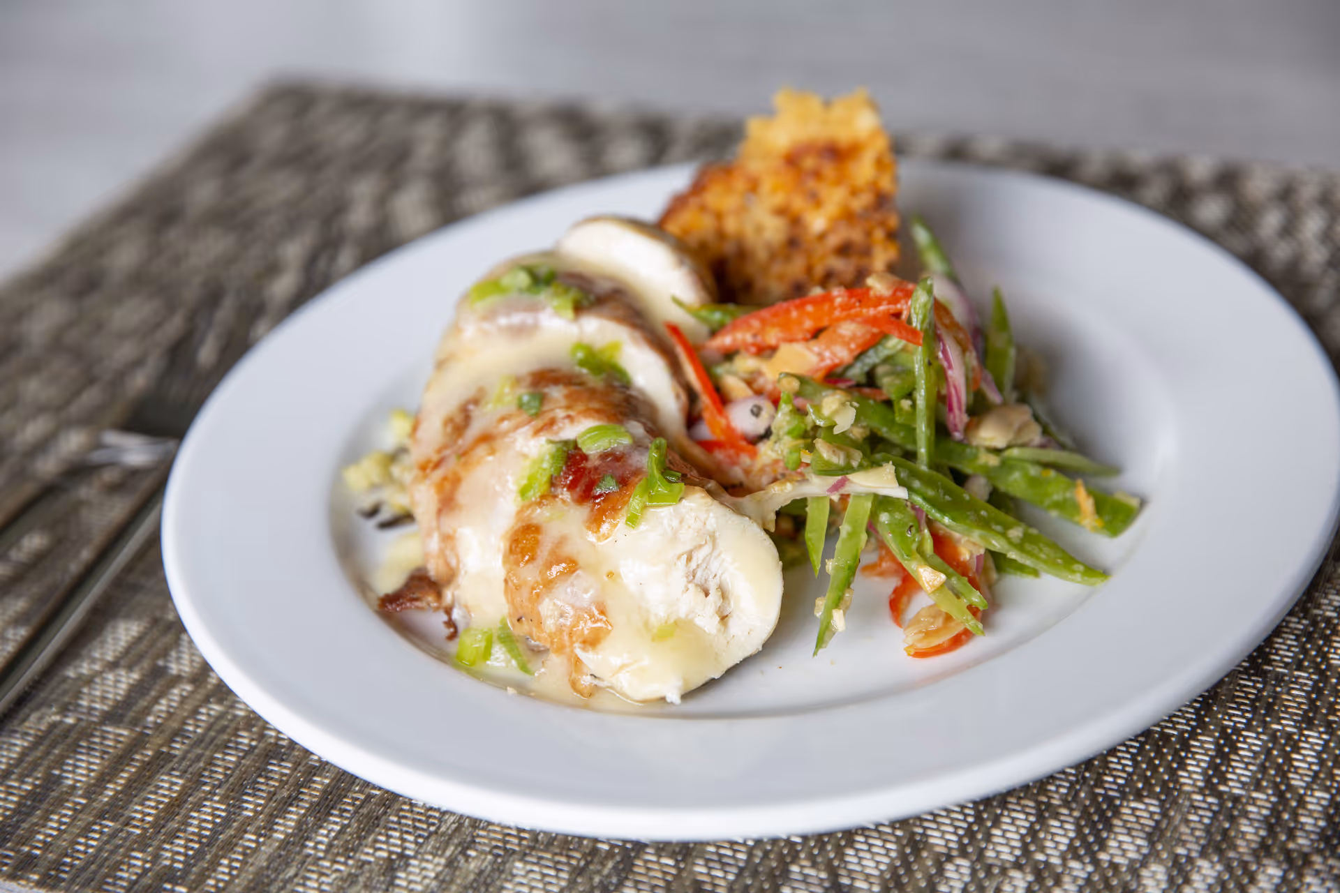 A white plate with sliced chicken breast topped with a creamy sauce and chopped green onions, served with a side of mixed vegetables including green beans and red bell peppers, and a piece of cornbread, placed on a woven placemat.