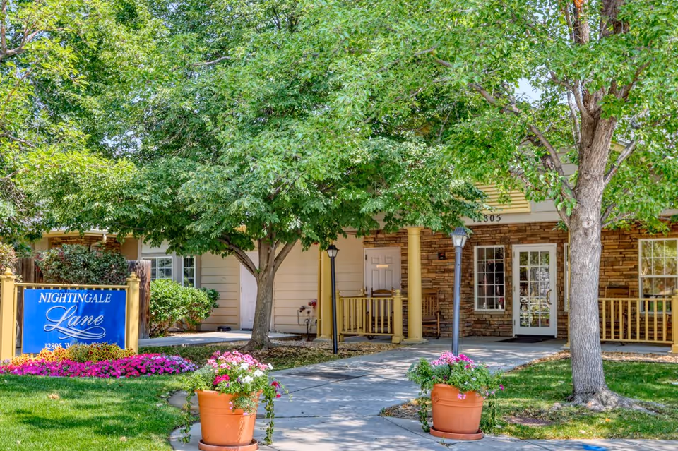 Exterior view of Nightingale Lane facility showing a pathway leading to the entrance surrounded by green trees and flower pots with pink and white flowers. The building has a stone facade with windows and a porch area with rocking chairs.