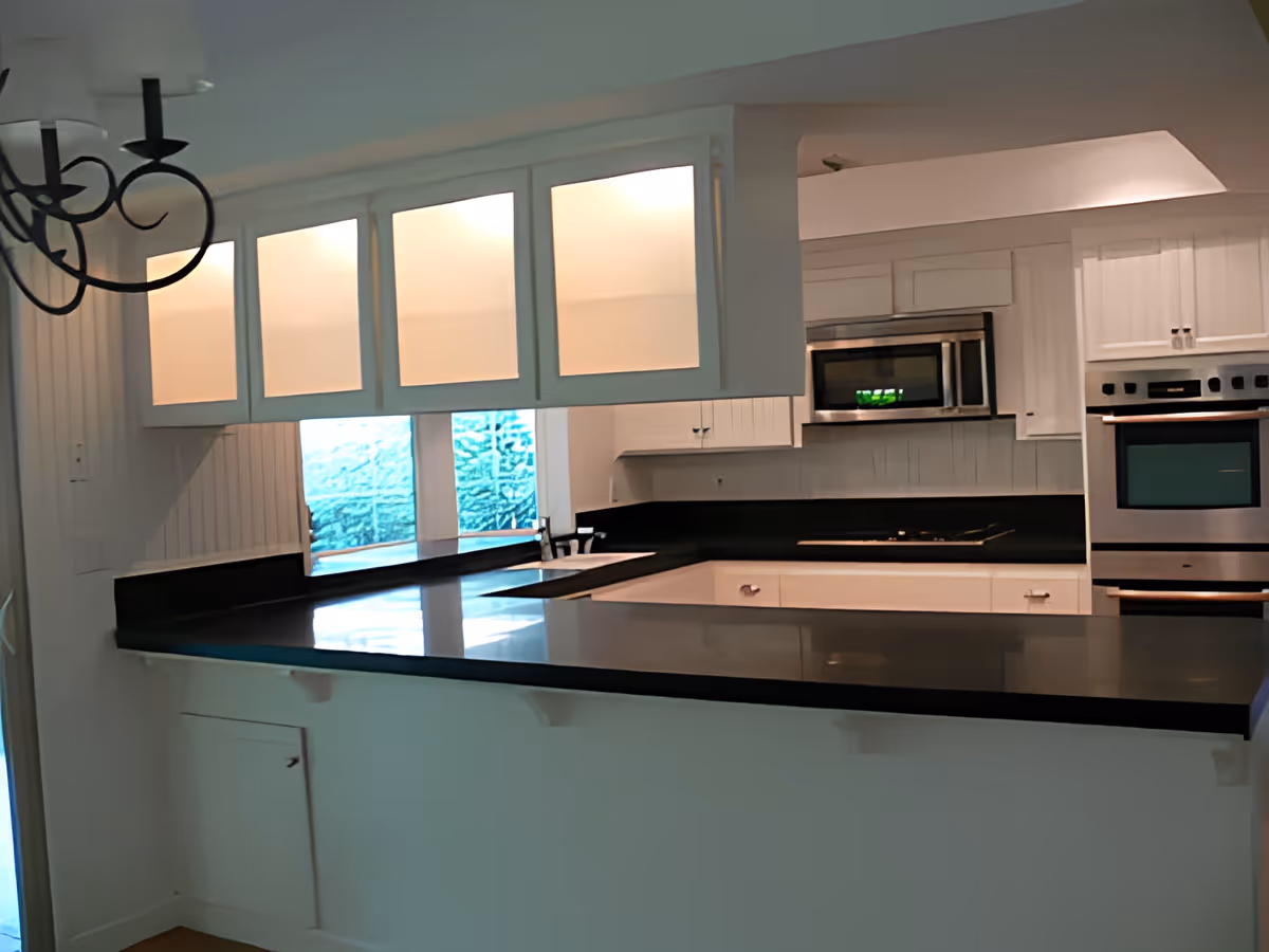 Modern kitchen with white cabinetry, black countertops, built-in stainless steel microwave and oven, and a black cooktop. There is a window above the sink and a decorative black chandelier hanging from the ceiling.