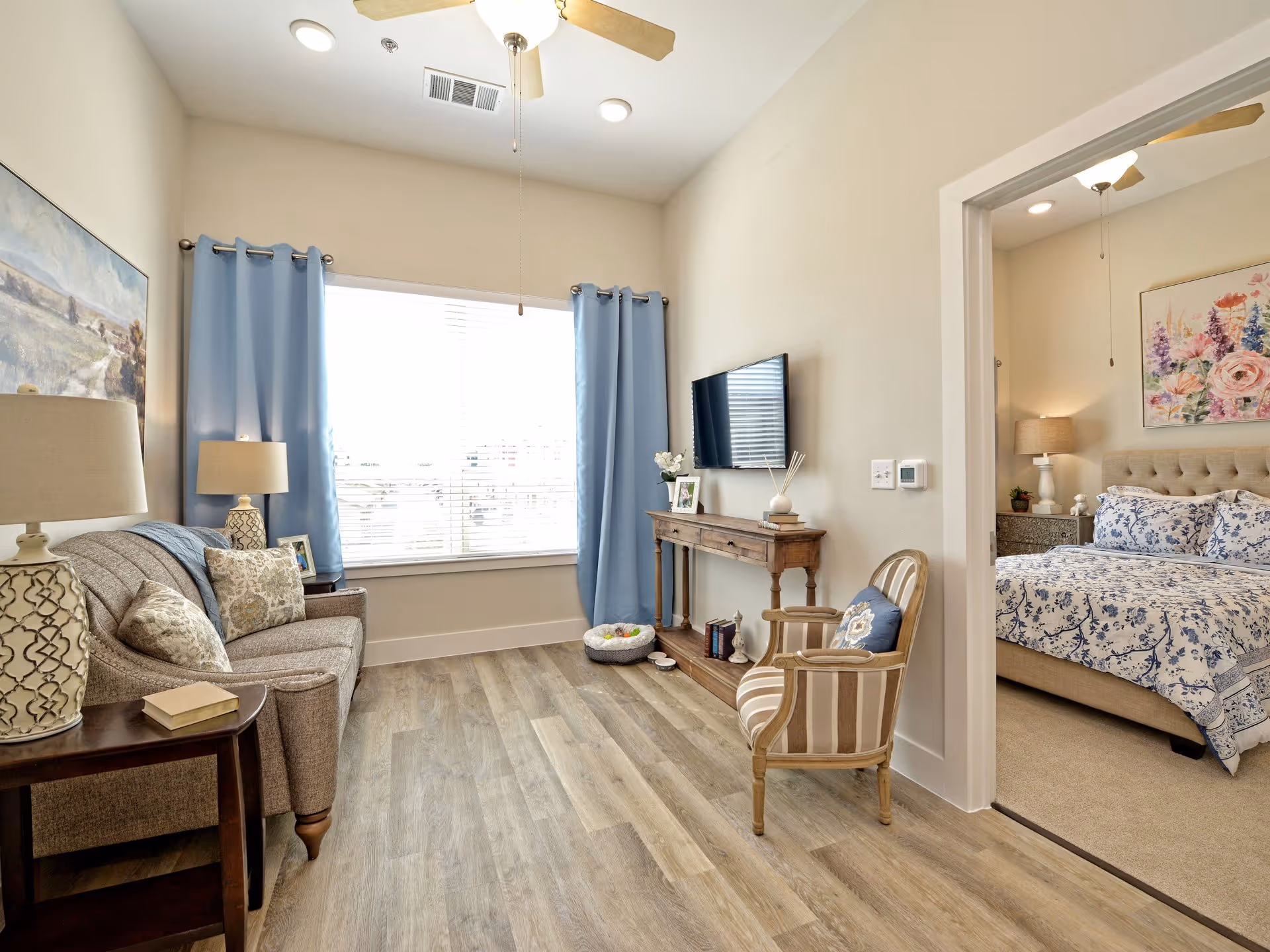 A bright living room with light wood flooring, a beige sofa with patterned cushions, a wooden side table with a lamp and book, a wooden console table with decorative items and a wall-mounted TV. Light blue curtains frame a large window. To the right, an open doorway leads to a bedroom with a bed dressed in blue and white floral bedding, a nightstand with a lamp, and a floral painting above the bed.