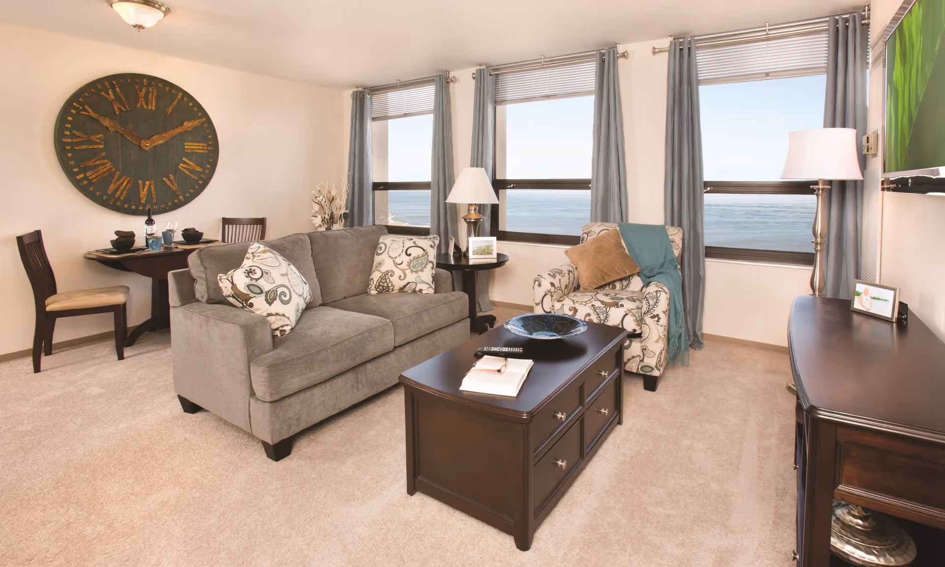 A cozy living room with a gray sofa adorned with patterned pillows, a matching armchair with a brown pillow and teal throw, a dark wooden coffee table with books and a decorative bowl, and a side table with a lamp and framed photo. Large windows with gray curtains offer a view of the ocean. A round wall clock with Roman numerals hangs above a small dining table with two chairs in the corner.