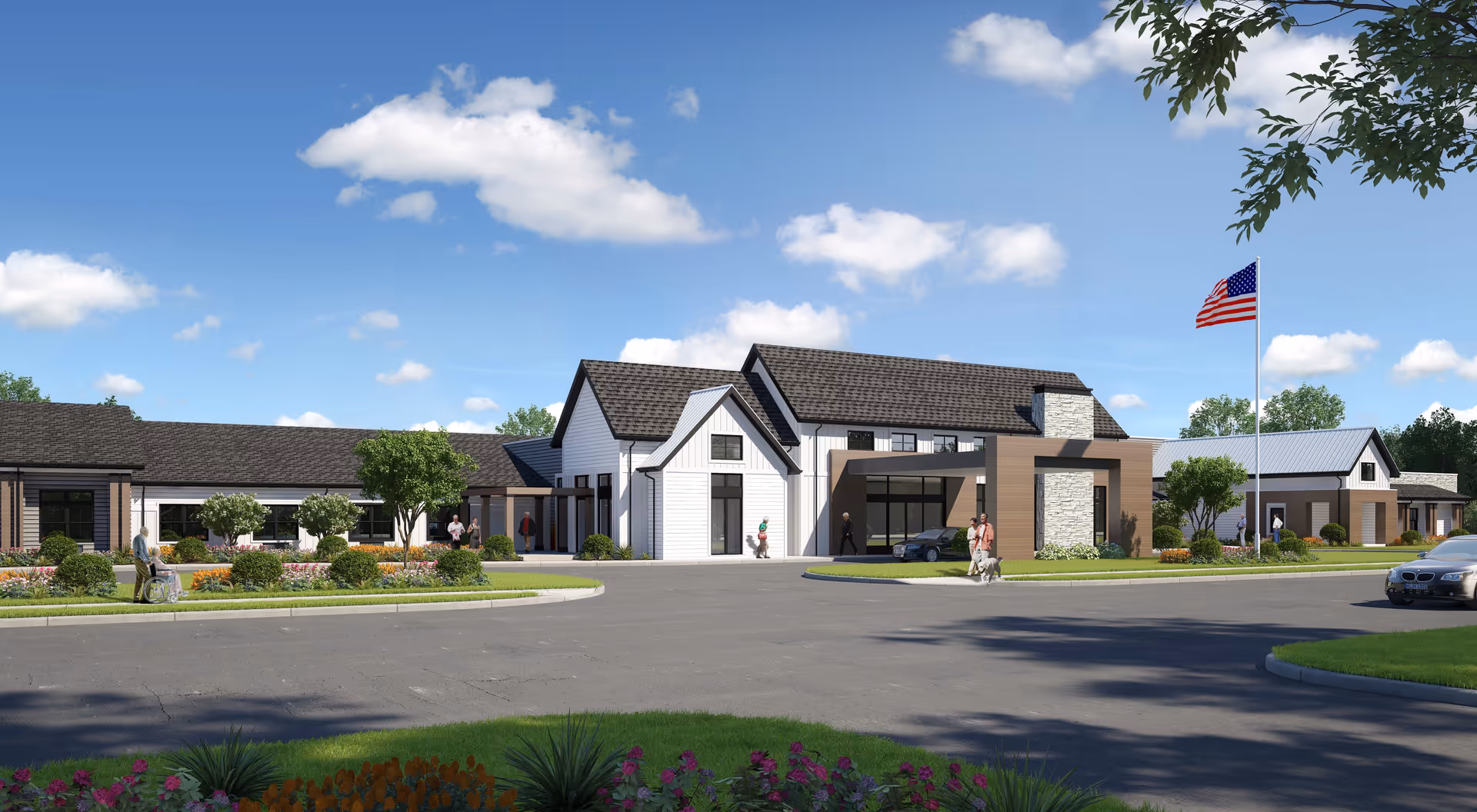 Exterior view of a modern senior living facility with a circular driveway, landscaped gardens, and an American flag on a flagpole. Several people are walking or sitting outside near the entrance under a clear blue sky with scattered clouds.