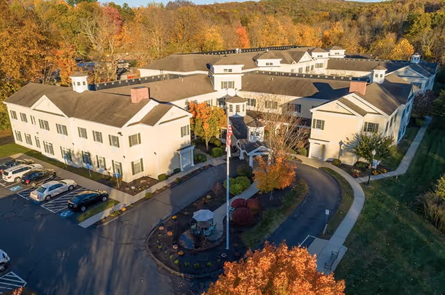 Aerial view of Monarch Southbury senior living facility surrounded by autumn trees with colorful foliage. The building is a large, light-colored, multi-wing structure with a circular driveway and landscaped garden area featuring a flagpole and outdoor seating. Several cars are parked in the adjacent parking lot.