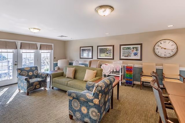 Bright senior living common room with a green sofa, patterned armchairs, tables and chairs, wall art, and a large clock near French doors.