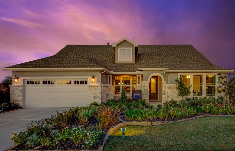 Front exterior of a single-story suburban house with an attached two-car garage, landscaped flowerbeds, and a purple sunset sky.