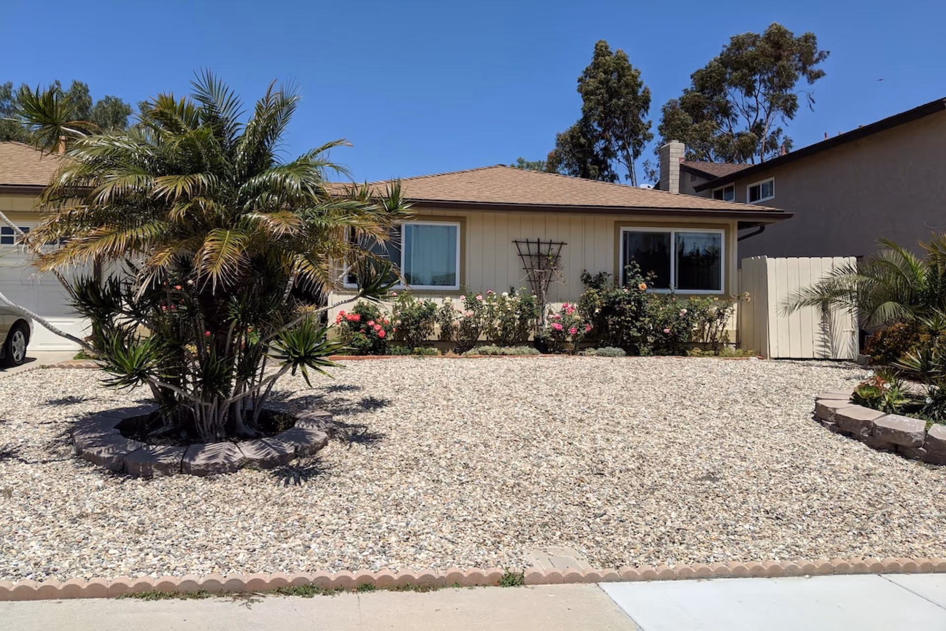 Front exterior view of a single-story house with a tan roof and beige siding. The front yard is covered with small rocks and features a circular planting area with a palm tree and other plants. There are two windows visible on the front of the house, and a white wooden gate on the right side. The sky is clear and blue.