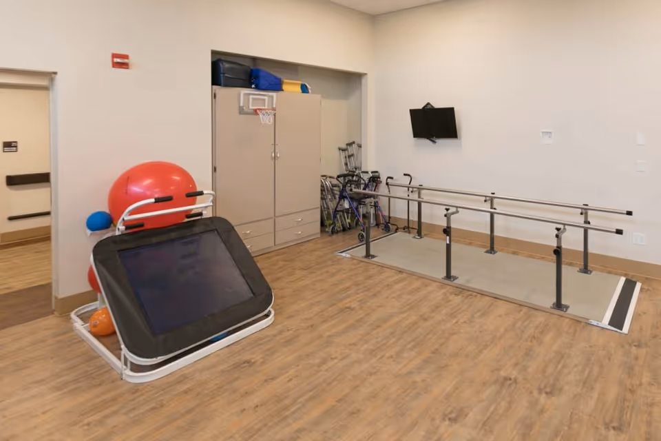 A rehabilitation room with wooden flooring featuring parallel bars for walking exercises, a wall-mounted TV, a cabinet with storage, and various physical therapy equipment including exercise balls, a trampoline, walkers, and crutches.