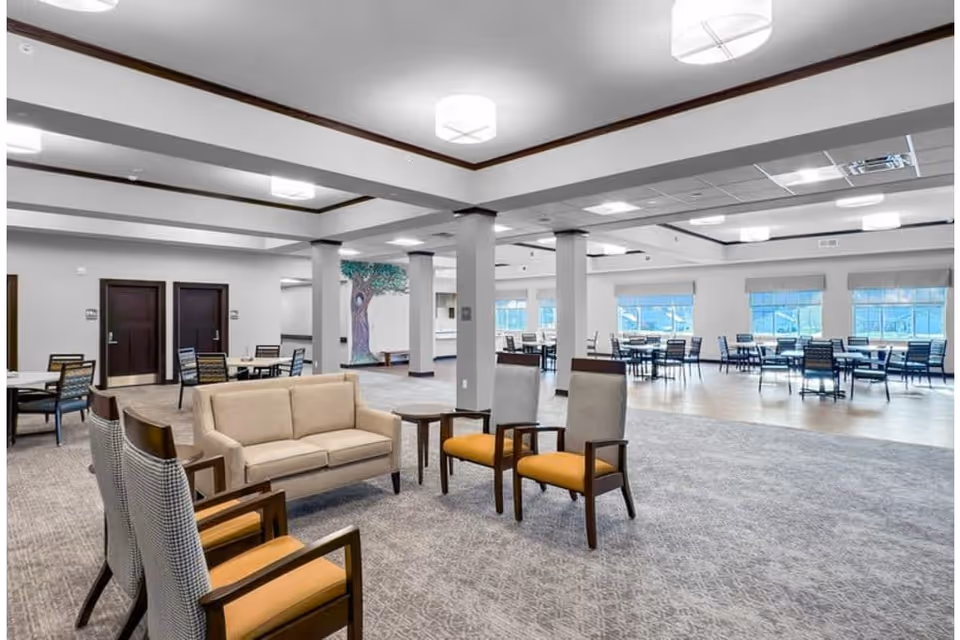 Spacious senior living common area with lounge seating in the foreground and multiple dining tables in the bright, open room.