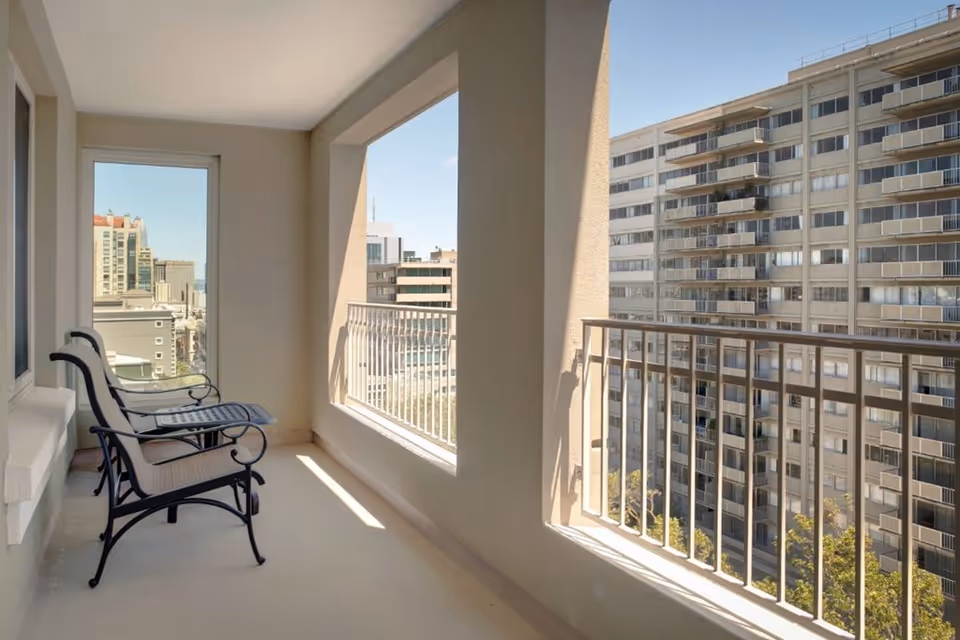 Covered balcony with two metal chairs and a small table overlooking neighboring apartment buildings.