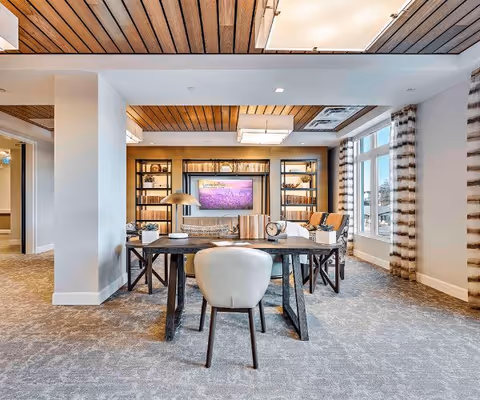 A bright and modern living room area with a wooden ceiling, large windows with striped curtains, and a carpeted floor. The room features a dark wooden table with a white chair in the foreground, a decorative lamp, and a wall-mounted TV flanked by shelves filled with books and decor items.