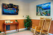 A cozy sitting area with two wooden rocking chairs, a small wooden table with books, a potted plant, a wall-mounted flat-screen TV displaying a colorful game, and a framed painting of a seaside village on the wall.