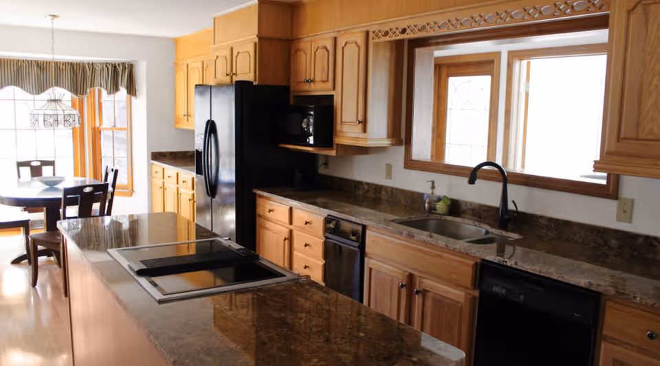 A kitchen with wooden cabinets, a black refrigerator, a built-in microwave, a sink with a black faucet, and a dishwasher. There is a large granite countertop island with a built-in stovetop. In the background, a dining area with a round table and chairs is visible near windows with a valance.