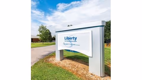 Freestanding entrance sign reading 'Liberty' for a care and rehabilitation center on a grassy lawn with trees and a blue sky.