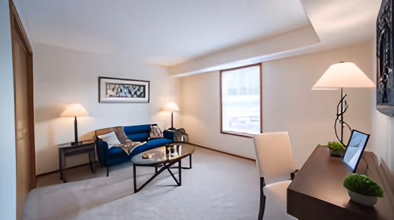A well-lit living room with a blue sofa, two side tables each with a lamp, a glass coffee table, a desk with a chair, a table lamp, and two small potted plants. There is a large window letting in natural light and a framed artwork on the wall above the sofa.