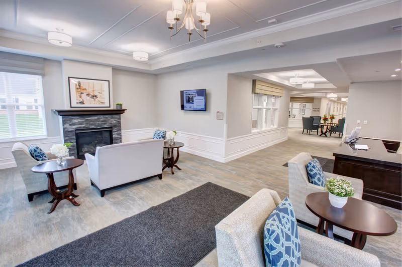 A bright and spacious senior living facility common area with comfortable seating including armchairs and sofas arranged around small wooden tables. There is a fireplace with a framed picture above it, a wall-mounted TV, and a reception desk to the right. The room has large windows with blinds, light-colored walls, and modern ceiling lights.