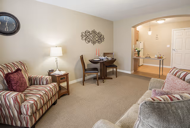 A furnished living room with striped armchairs, a small round dining table, a table lamp, wall decor, and an archway opening to a kitchenette.
