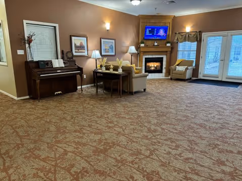 A spacious living room area with patterned carpet, a wooden piano against the left wall, a small table with two chairs, two armchairs near a fireplace with a mounted TV above it, and double glass doors with windows on the right side letting in natural light.