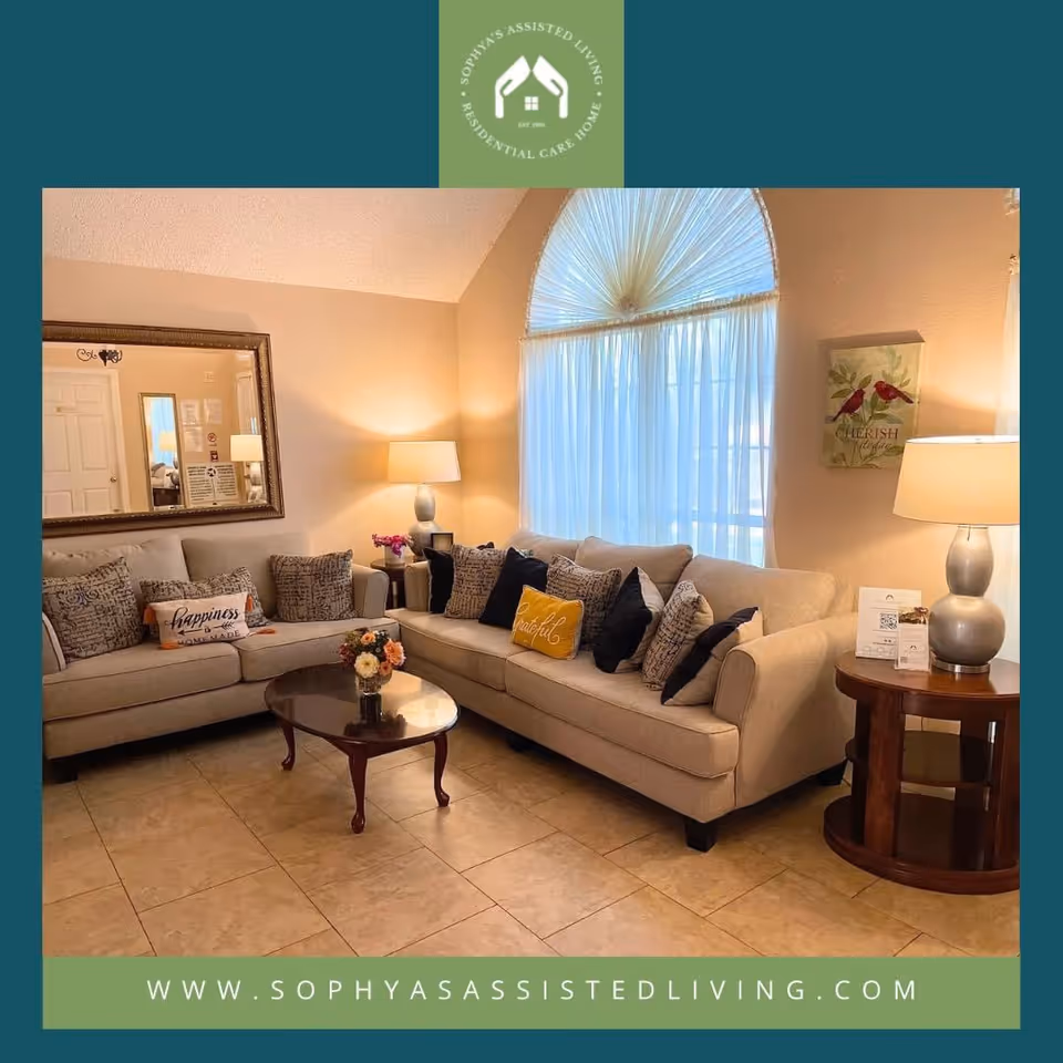 A cozy living room with two beige sofas adorned with multiple decorative pillows, a wooden coffee table with a small flower arrangement, two table lamps on side tables, a large mirror on the wall, and a large window with sheer curtains allowing natural light to fill the room.