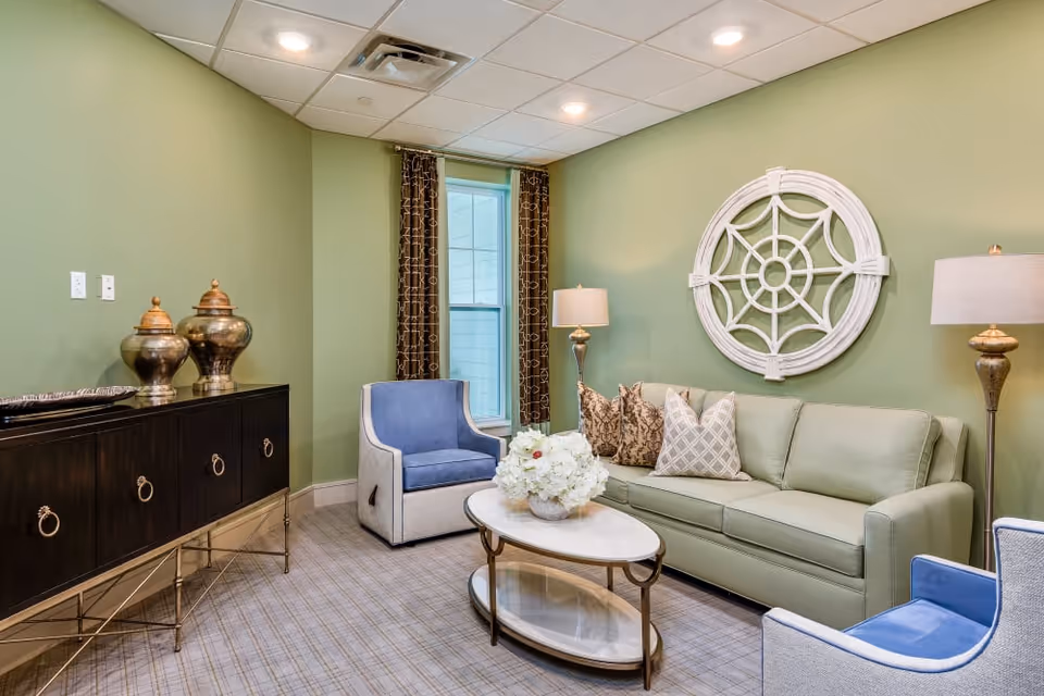 A cozy living room with light green walls, a light green sofa adorned with patterned pillows, two blue and white armchairs, a white oval coffee table with a floral centerpiece, a dark wooden sideboard with decorative vases, two floor lamps, and a large white circular wall decoration resembling a window frame. A window with patterned curtains is visible in the background.