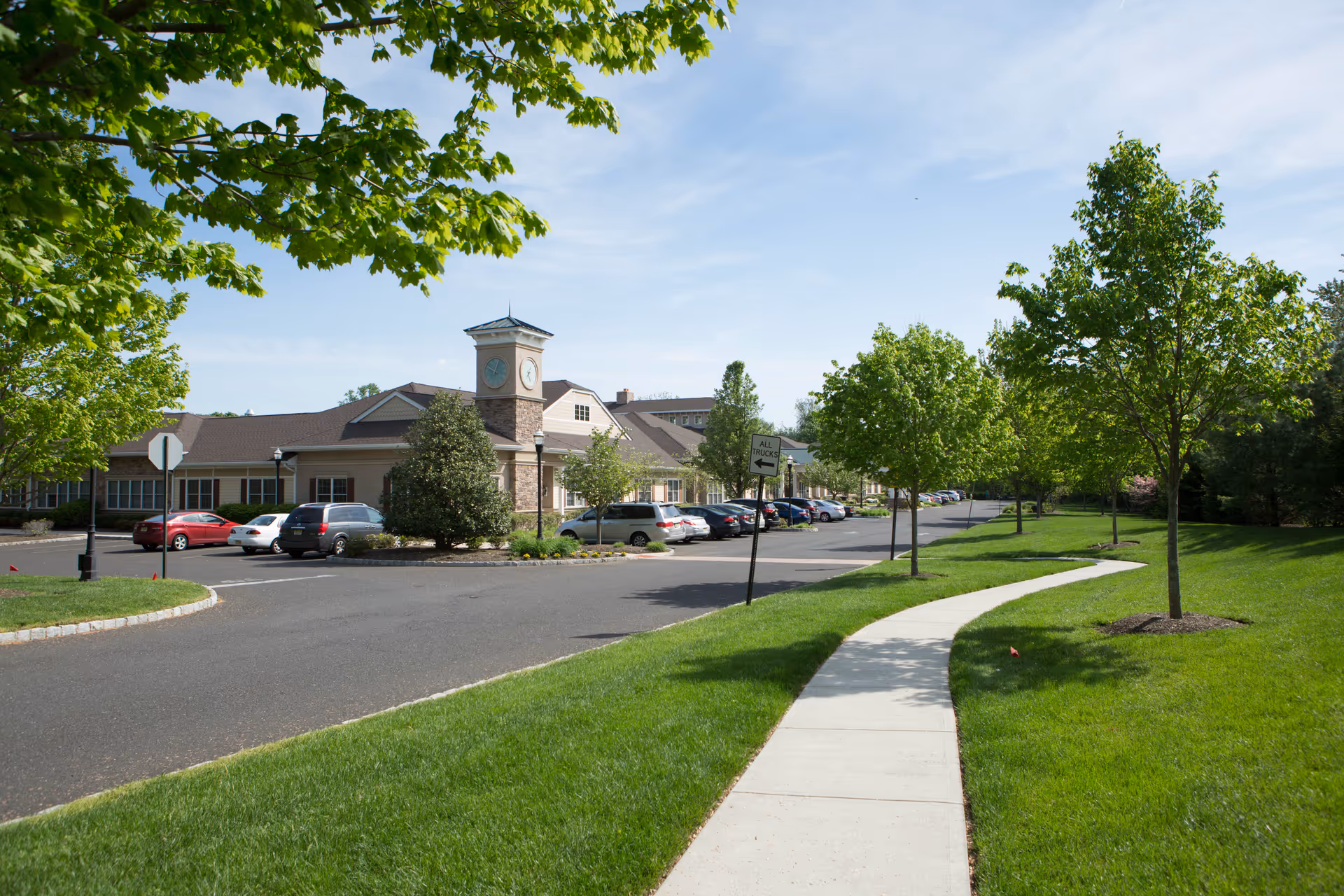 View of the exterior of Harmony Village at Stanwick Road facility on a sunny day, showing a paved driveway with parked cars, a clock tower on the building, green trees, and a curved sidewalk with well-maintained grass.