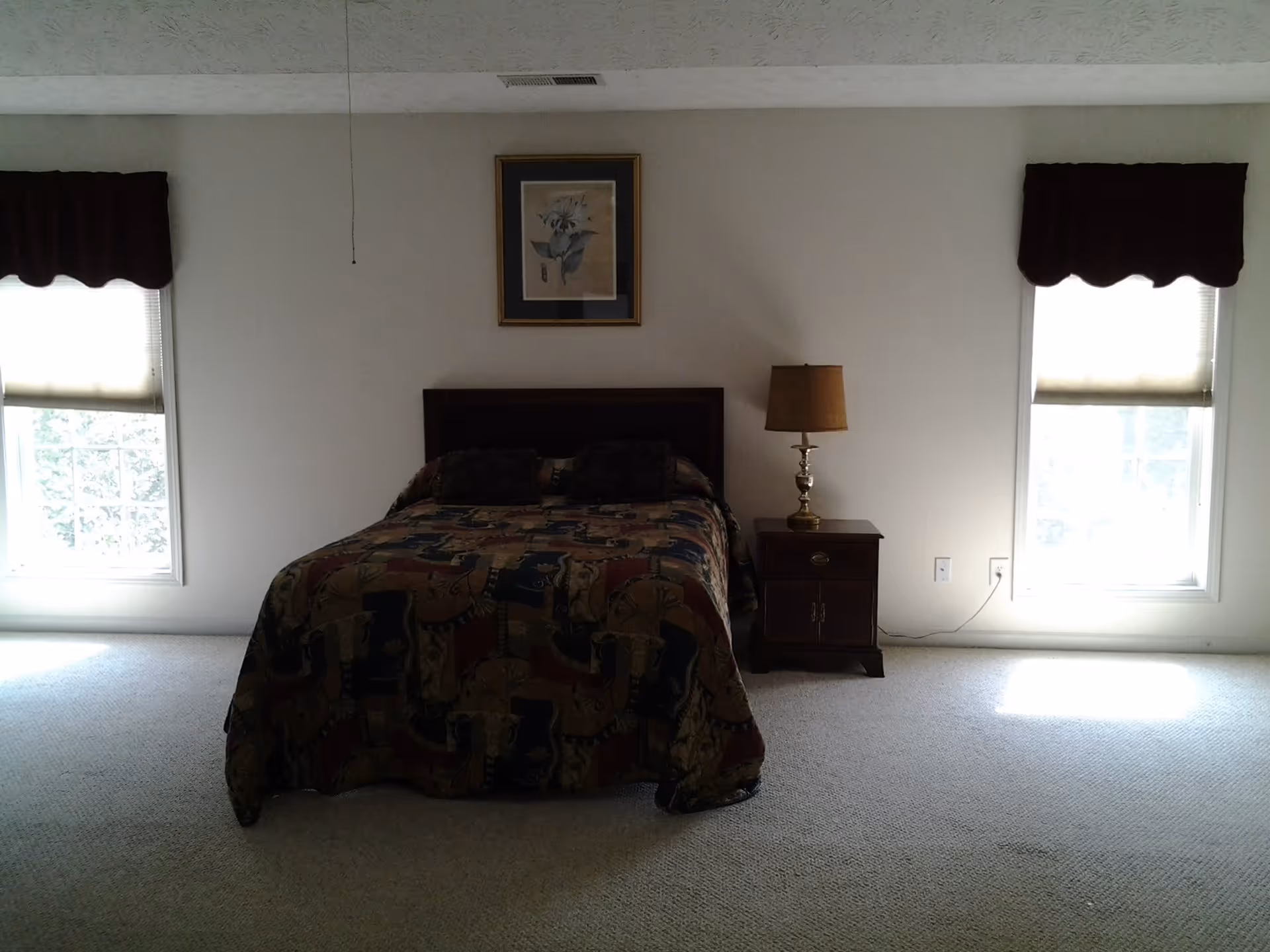 A sparsely furnished bedroom with a double bed centered between two windows and a nightstand with a lamp.
