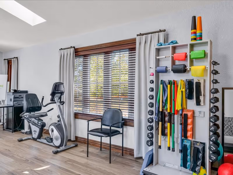 A bright room with exercise equipment including a stationary recumbent bike, a black chair, and a wall-mounted rack holding various colorful exercise bands, weights, and cones. The room has large windows with wooden blinds and light-colored curtains, and a wooden floor.
