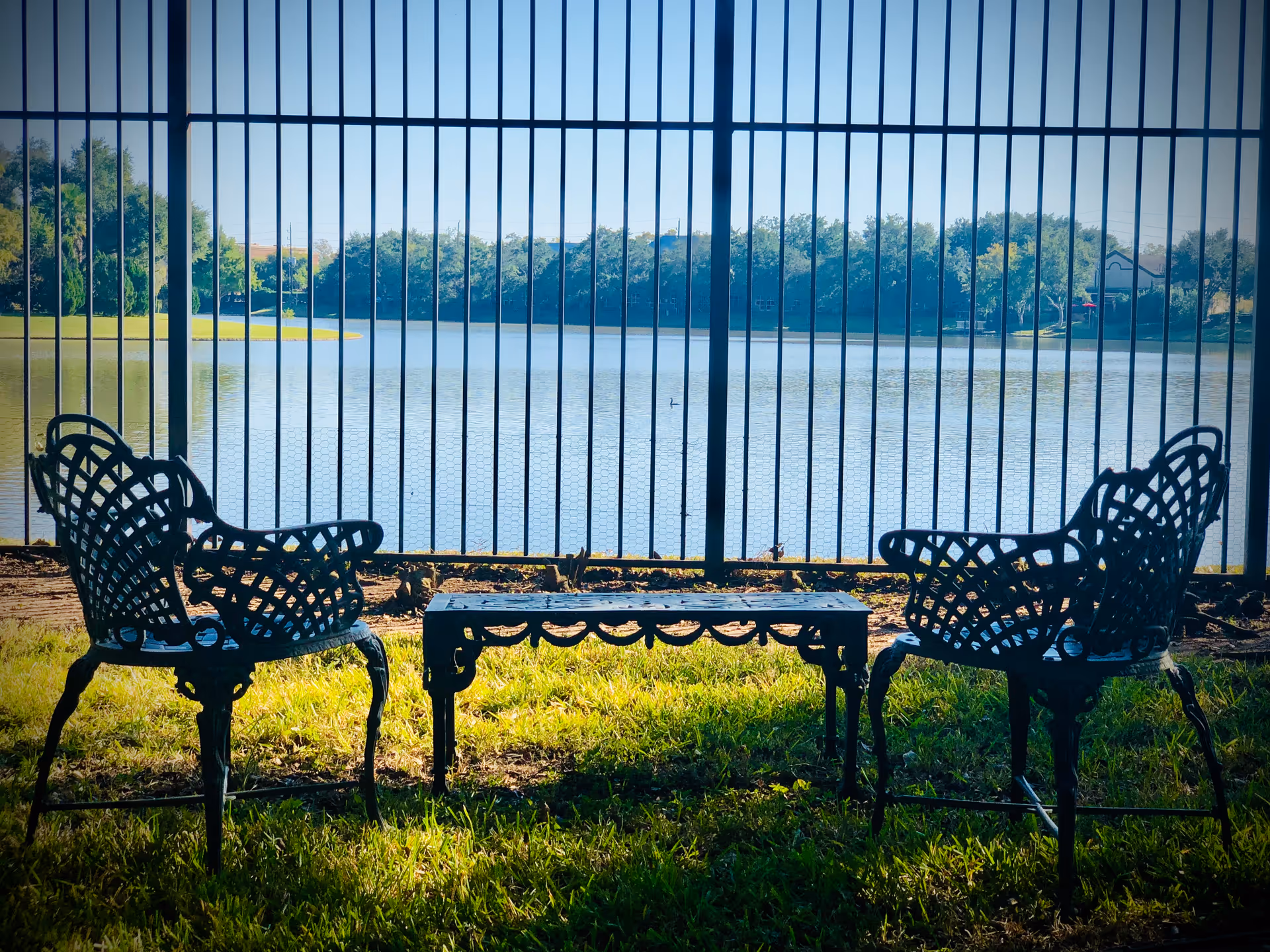 Two ornate metal chairs and a matching metal table on grass, positioned in front of a black metal fence with a view of a lake and trees in the background under a clear sky.