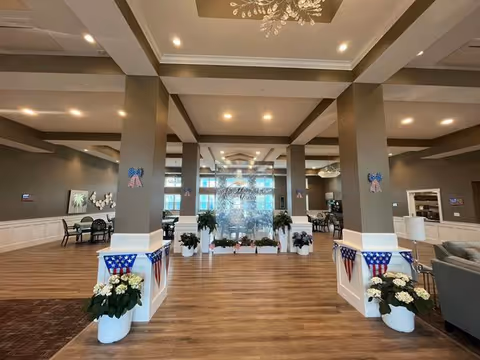 Spacious and well-lit interior common area of an assisted living facility with wooden flooring, large columns decorated with patriotic ribbons and banners, potted plants, and seating areas with tables and chairs. A glass wall with the facility name 'Hampton Manor' is visible in the center background.