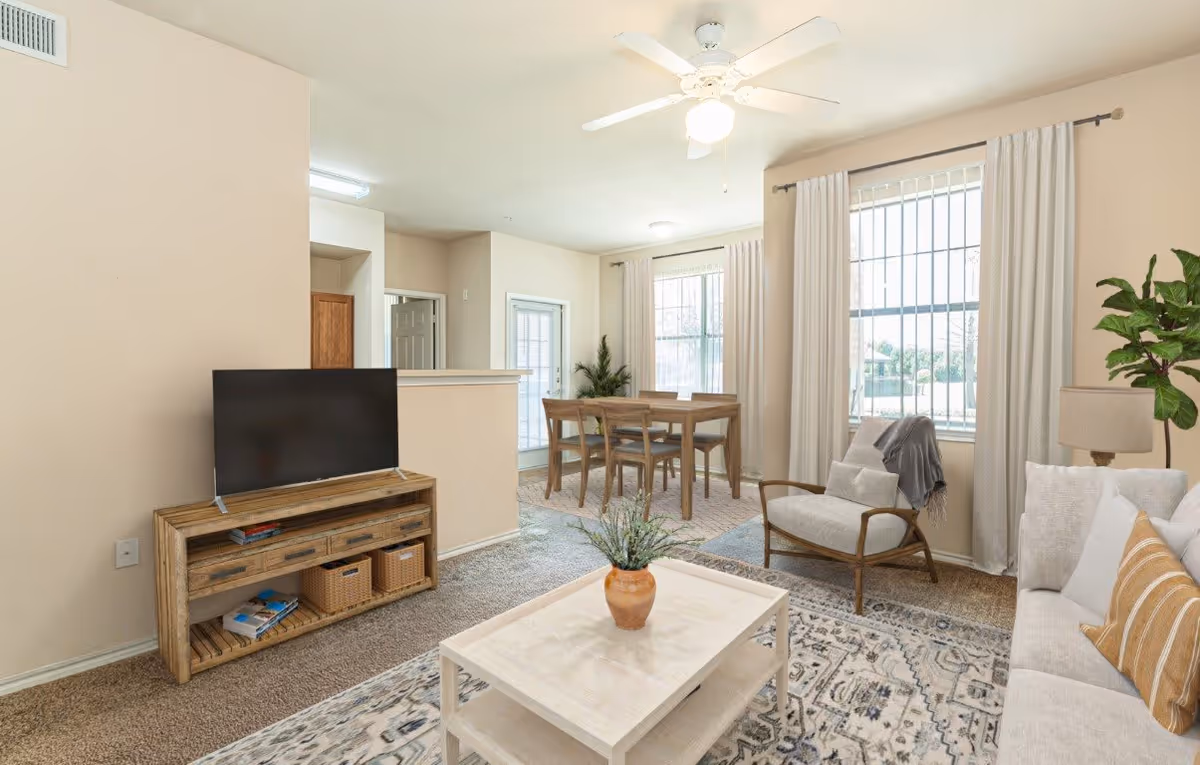 A bright living room and dining area in an apartment with beige walls and carpeted floors. The living room features a light-colored sofa with pillows, a wooden armchair with a throw blanket, a coffee table with a small plant, and a wooden TV stand with a flat-screen TV. The dining area has a wooden table with four chairs, two large windows with white curtains, and a ceiling fan with a light fixture above.