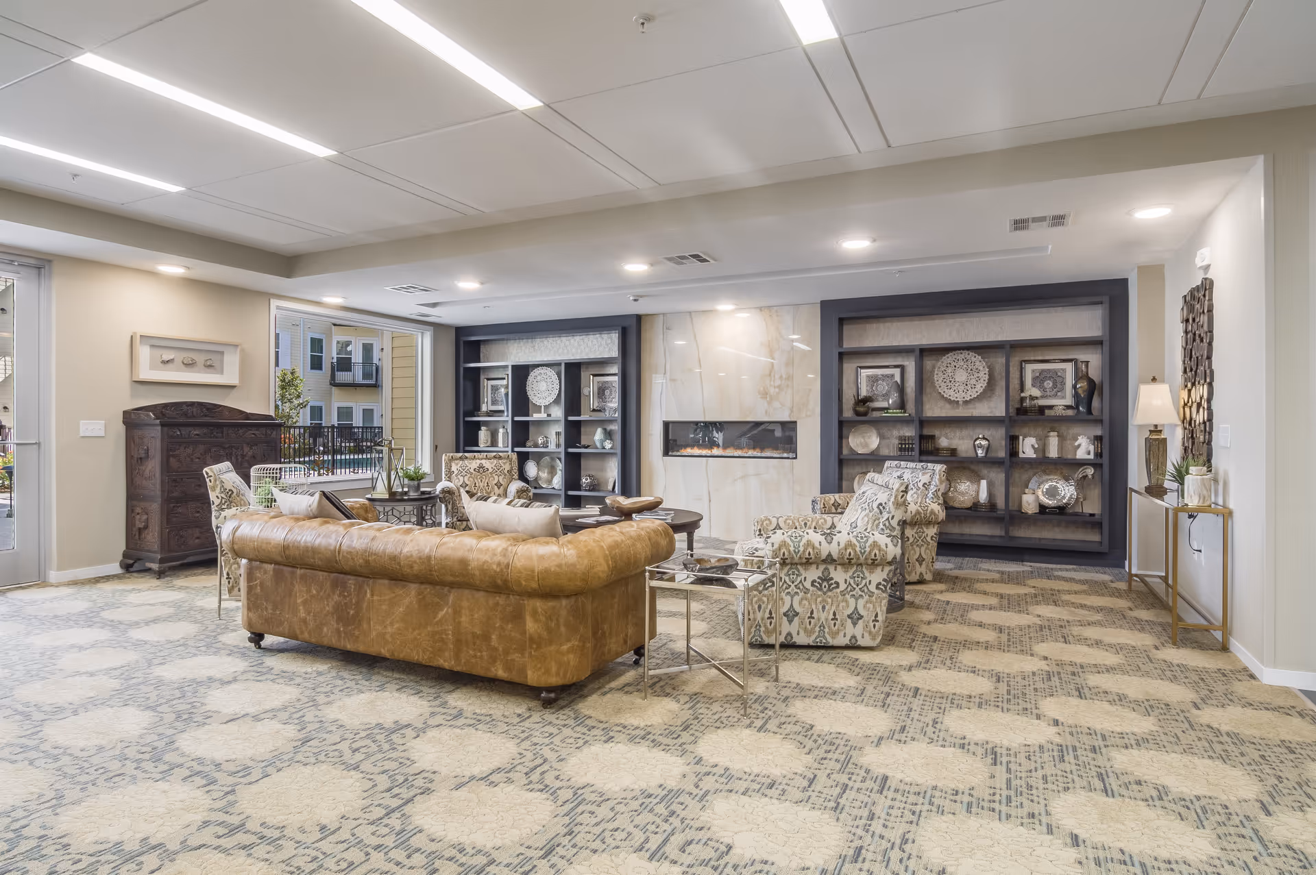 A spacious and well-lit living room area with a brown leather sofa, patterned armchairs, a modern fireplace set in a marble wall, and built-in shelves displaying decorative plates and vases. The room has a patterned carpet and neutral-colored walls, with a door leading outside and a small table with a lamp on the right side.