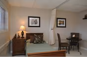 A small bedroom with a single bed featuring a green bedspread and decorative pillows. Next to the bed is a wooden nightstand with a lamp and some decorative items. Two framed pictures hang on the beige walls. There is a small round table with two chairs in the corner, and a white privacy curtain partially drawn between the bed and the table. The room has a window with blinds and a tiled ceiling.