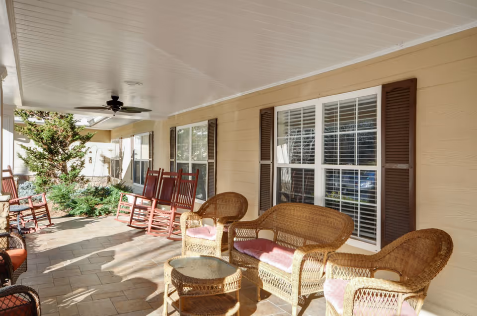 Covered outdoor patio area with wicker chairs and a loveseat with pink cushions, a glass-top wicker table, and several wooden rocking chairs. The patio has a ceiling fan and is adjacent to a beige building with windows and brown shutters. There are some green shrubs and plants near the edge of the patio.