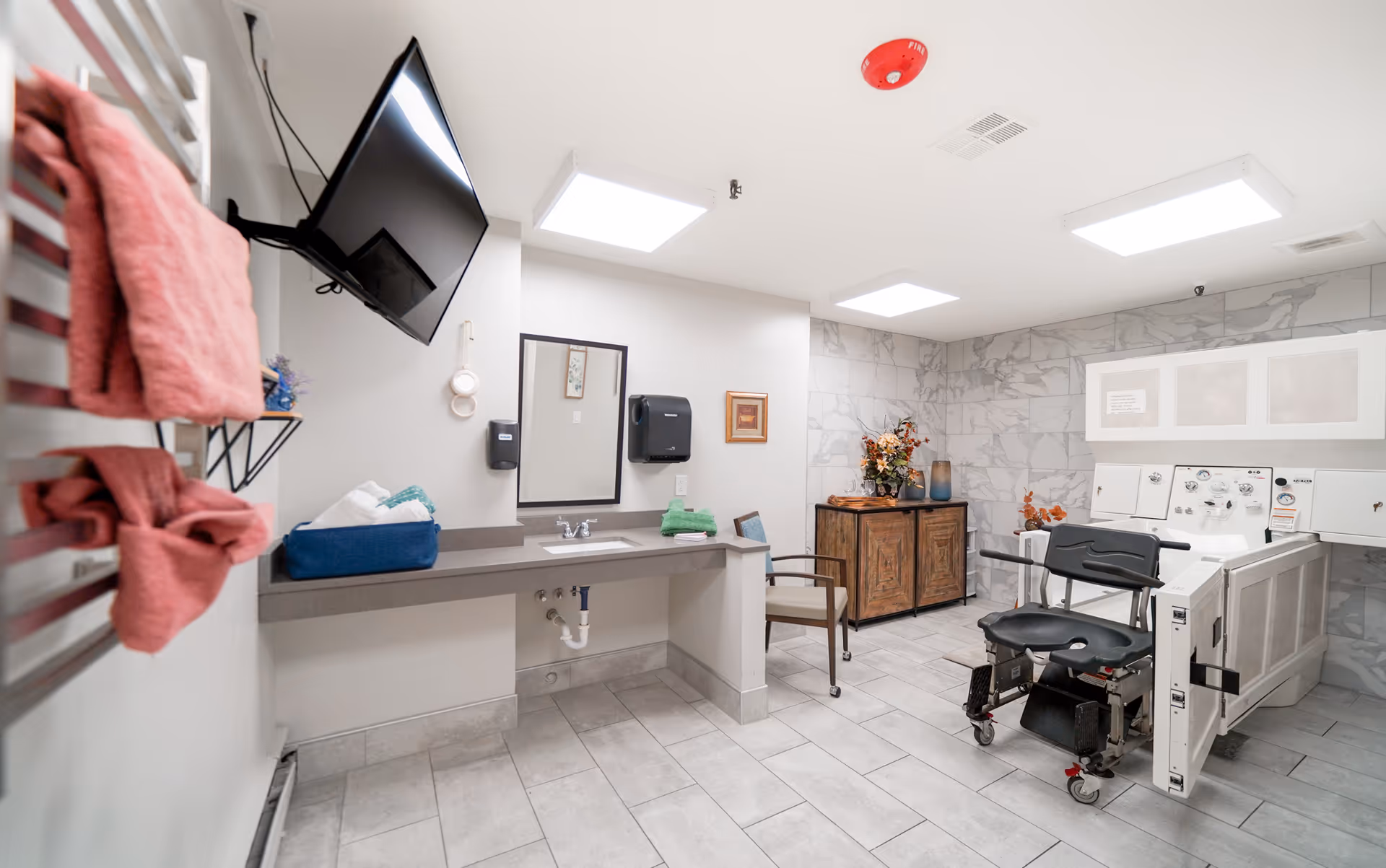 A bright, clean room with tiled floors and walls, featuring a mounted flat-screen TV, a countertop with a sink, a mirror, and a paper towel dispenser. There are pink towels hanging on a rack, a chair, a wooden cabinet with decorative items, and a specialized bathing chair next to a large accessible bathtub.